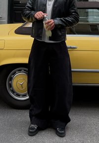 Fekete, széles szárú nadrág texturált anyagból, szürke cipzáros pulóverrel és fekete bőrdzsekivel párosítva, kezében átlátszó italos palack.