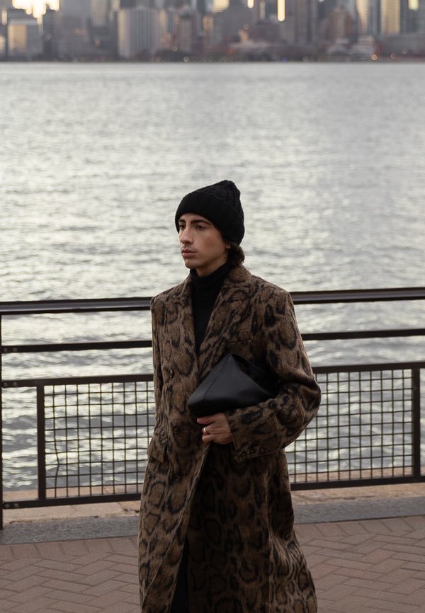Homme en manteau à motif léopard et bonnet noir tenant une pochette noire, debout au bord de l'eau avec la ligne d'horizon de la ville en arrière-plan au crépuscule.