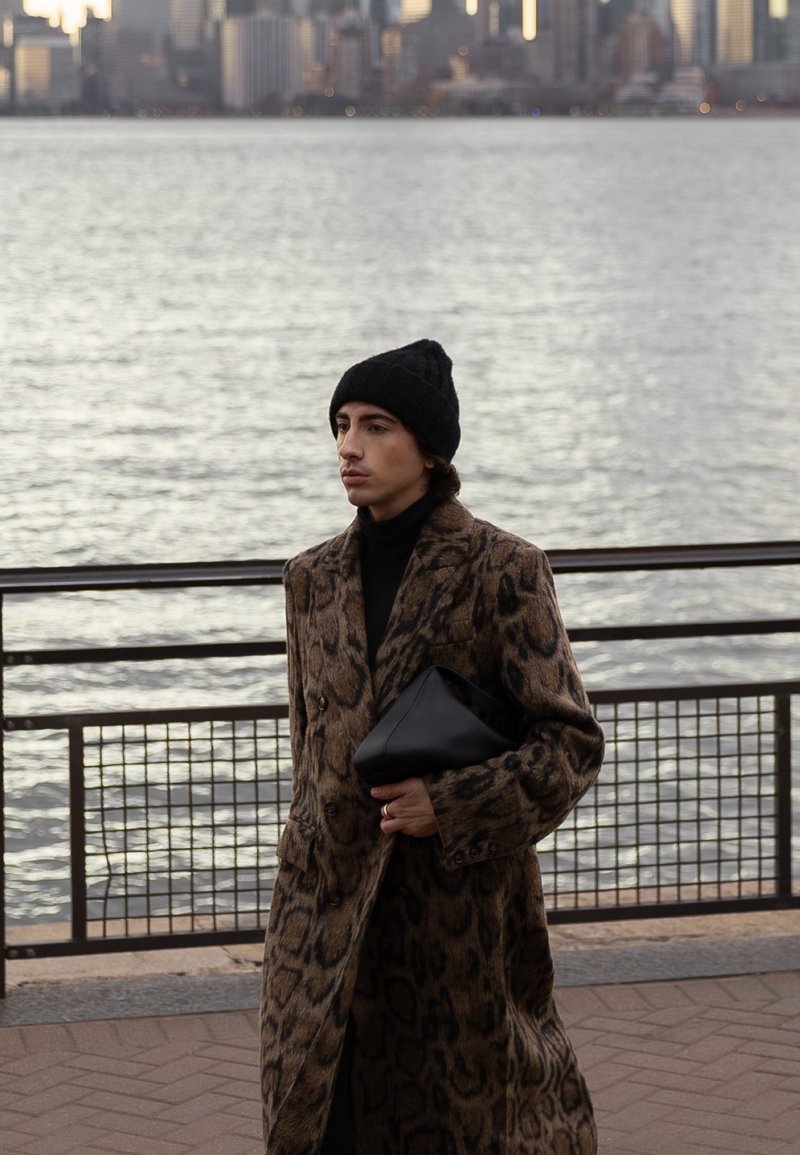 Homme en manteau à imprimé léopard et bonnet noir tenant une pochette noire, debout au bord de l'eau avec la silhouette d'une ville en arrière-plan au crépuscule.