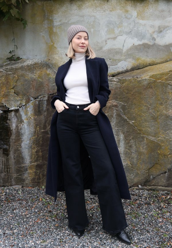 Una mujer con un gorro de punto gris, una prenda superior blanca, un abrigo largo azul marino y pantalones negros posa junto a un muro de piedra desgastado.