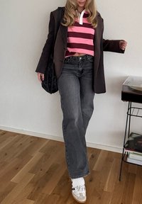 Modèle portant un blazer marron sur un polo à rayures roses, associé à un jean large gris et des baskets blanches avec des accents gris, debout à l'intérieur.