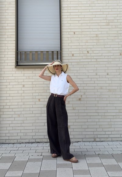 Mujer con sombrero de ala ancha, camiseta sin mangas blanca y pantalones negros sueltos, está apoyada contra una pared clara de ladrillo cerca de una ventana cerrada.