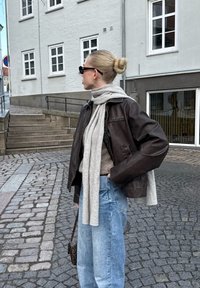 Bruine leren jas over een grijze trui, gecombineerd met lichtblauwe wijde jeans. Gewikkeld in een grijze sjaal, geaccessorieerd met zwarte zonnebril.