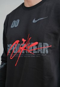 Sweatshirt noir avec un logo Nike, un texte graphique rouge audacieux et des noms de villes en gris. Manches longues avec des accents à motifs. Matière en coton.
