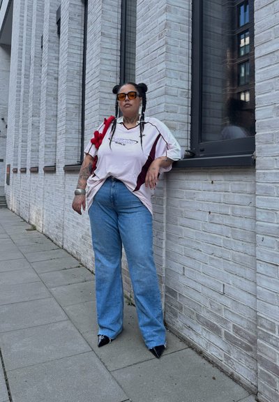 Camisa deportiva de gran tamaño en rosa claro y burdeos, jeans azul claro, tacones negros de punta, bolso rojo con pompones, peinado trenzado, entorno urbano.