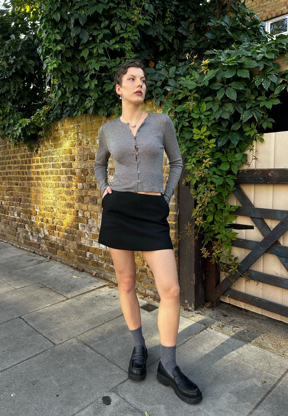 Γκρι μακρυμάνικη κάρντιγκαν με κούμπωμα, συνδυασμένη με μαύρη μίνι φούστα. Μαύρες λοφάρες με γκρι κάλτσες, τοποθετημένες μπροστά σε τοίχο από τούβλα και ανάγκη.