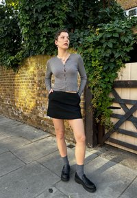 Γκρι μακρυμάνικη κάρντιγκαν με κούμπωμα, συνδυασμένη με μαύρη μίνι φούστα. Μαύρες λοφάρες με γκρι κάλτσες, τοποθετημένες μπροστά σε τοίχο από τούβλα και ανάγκη.