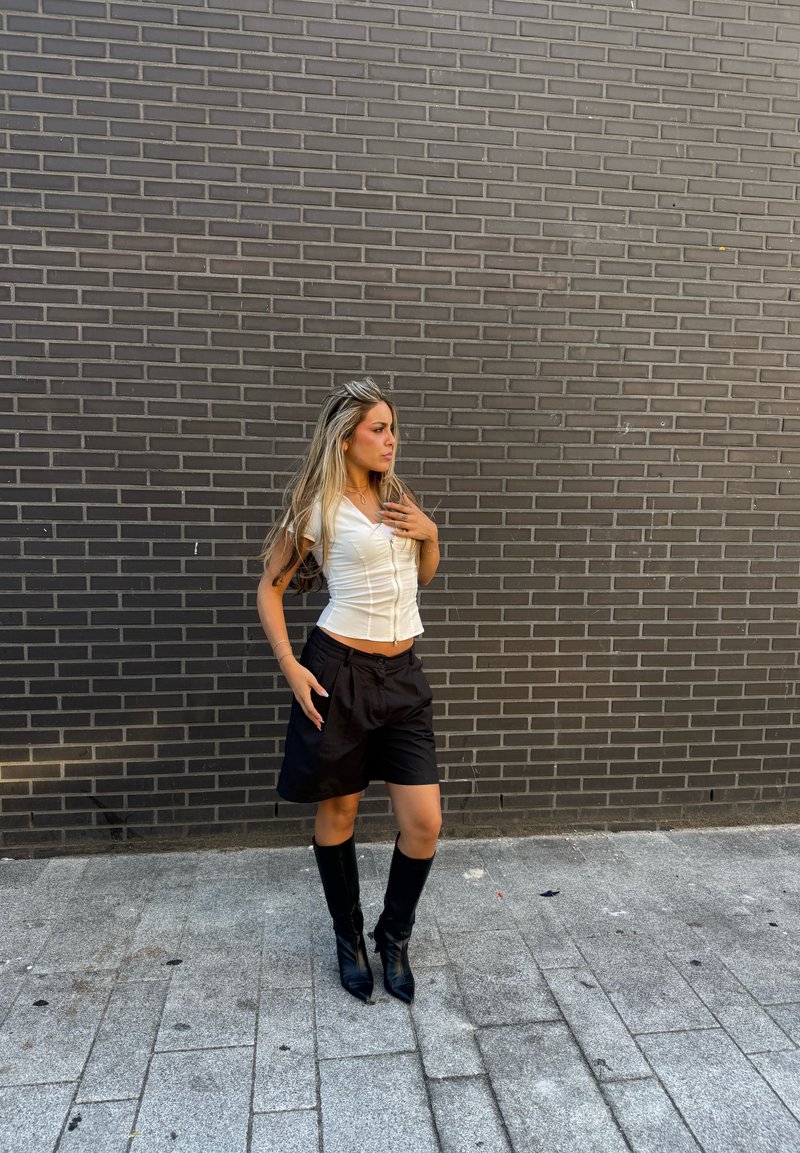 Giovane donna con capelli lunghi che indossa una canotta bianca, pantaloncini neri e stivali neri al ginocchio, in piedi contro un muro di mattoni grigio scuro.