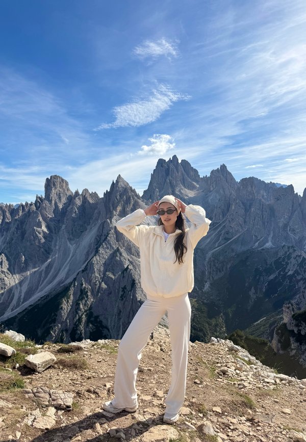 Woman in white outfit and sunglasses poses on rocky mountain trail with jagged peaks and blue sky in the background.