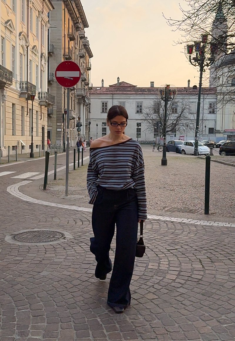 Žena s okuliarmi, pruhovaným topom s odhalenými ramenami a čiernymi nohavicami kráča po kamennej dlažbe s budovami a autami v pozadí pri západe slnka.