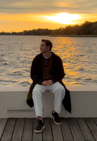 Joven sentado en un saliente junto al agua, vistiendo un abrigo oscuro sobre un suéter marrón, pantalones blancos y zapatillas oscuras. De fondo, se aprecia un atardecer.