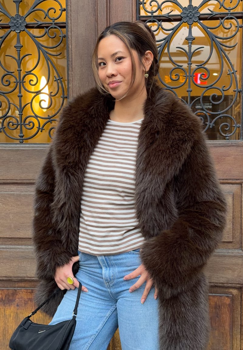 Cappotto marrone in pelliccia sintetica con texture soffice, indossato sopra una maglia a righe. Jeans azzurri e una borsa nera completano l'outfit.