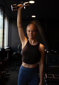 Zwarte dumbbell boven het hoofd gehouden, met een kort zwart tanktop en blauwe high-waisted leggings. De achtergrond toont een donkere gymomgeving.