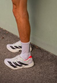 Nogi w białych butach sportowych z czarnymi paskami i grubych białych skarpetach stoją na teksturowanej nawierzchni obok jasnoszarej ściany.
