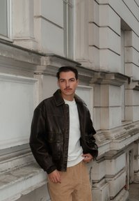 Chaqueta de cuero marrón con cremallera frontal, que cuenta con dos bolsillos en el pecho, usada sobre una camisa blanca. La persona lleva puesto un pantalón de pana beige.