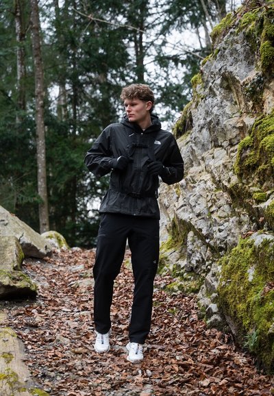 Chaqueta negra impermeable con capucha y puños ajustables, combinada con pantalones negros y zapatos blancos, de pie entre rocas y hojas caídas.