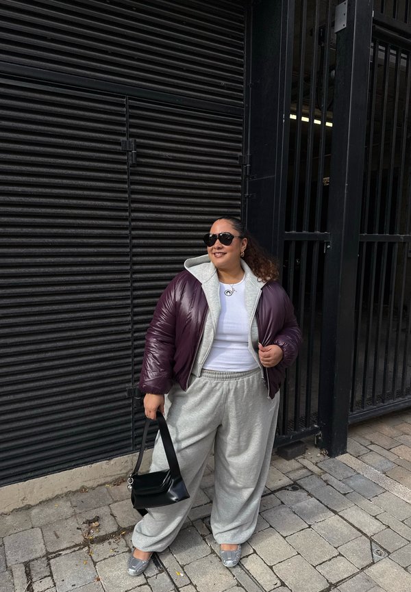 Una mujer con gafas de sol, una chaqueta color ciruela y pantalones de chándal grises está de pie en una acera de la ciudad y sostiene un bolso negro.