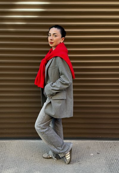 Blazer gris a medida sobre pantalones gris claro, con una bufanda de punto roja drapeada alrededor del cuello. El fondo presenta rayas horizontales marrones.