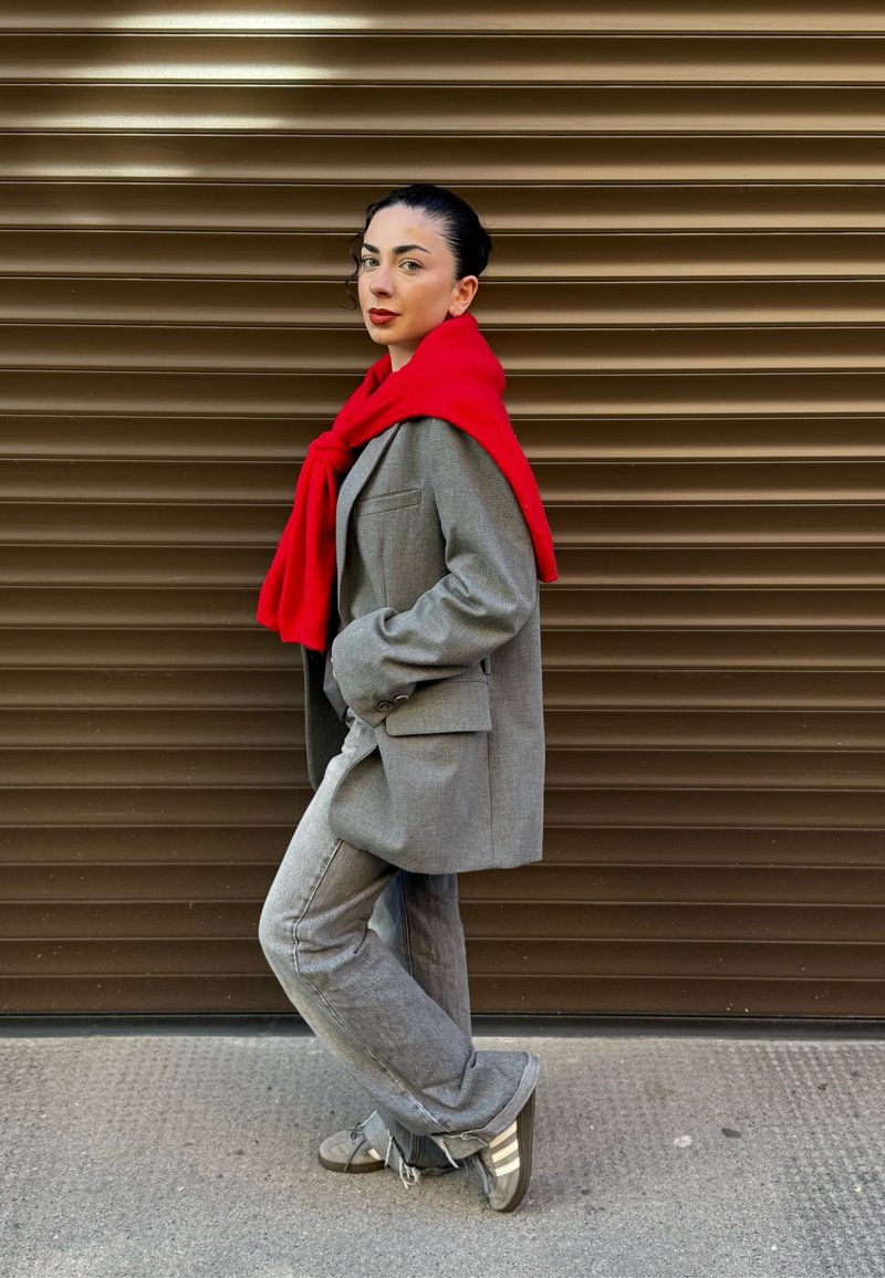 Blazer gris ajusté sur un pantalon gris clair, avec une écharpe tricotée rouge drapée autour du cou. L'arrière-plan présente des rayures horizontales marron.