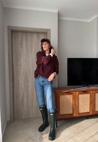 Femme portant un pull bordeaux, un jean, des bottes de pluie vertes et une casquette, debout à l'intérieur près d'une télévision posée sur un meuble en bois.