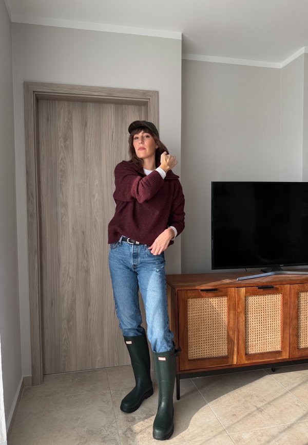 Žena u bordo džemperu, trapericama, zelenim čizmama za kišu i kapom, stoji unutra blizu televizora na drvenom ormaru.