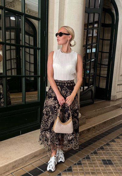 Blusa blanca sin mangas con pliegues verticales, falda negra con patrones dorados ornamentales, bolso beige y zapatillas blancas.