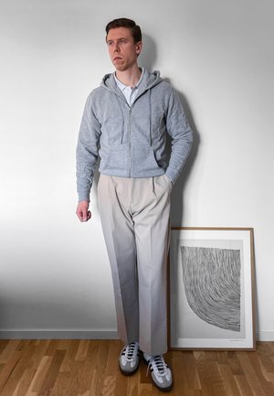Hombre con sudadera gris con cremallera, pantalones beige claros y zapatillas de deporte, de pie junto a una obra de arte abstracta en blanco y negro enmarcada, sobre un suelo de madera.