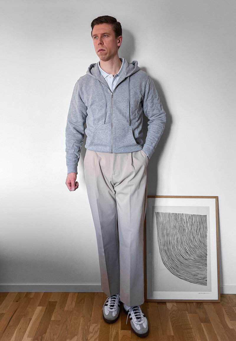 Bărbat îmbrăcat într-un hanorac gri cu fermoar, pantaloni bej deschis și adidași, stând lângă o operă de artă abstractă alb-negru, înrămată, pe podea de lemn.