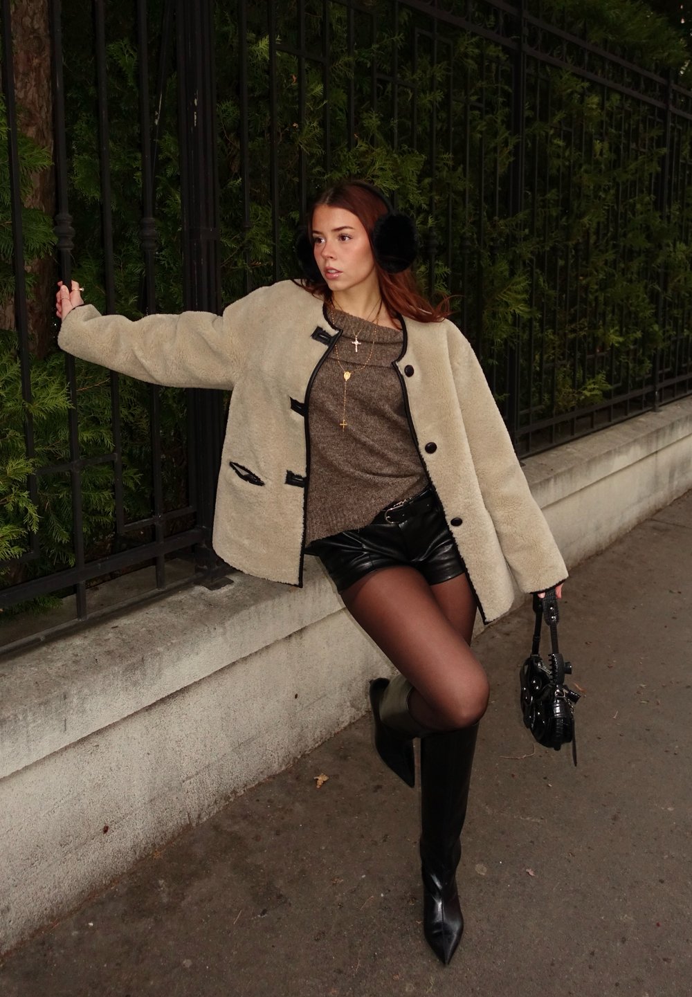 Beige gyapjúpelerő kabát fekete díszítéssel, barna pulóver, fekete bőr sort, áttetsző harisnyanadrág, térd fölé érő fekete csizma, és kis fekete kézitáska.