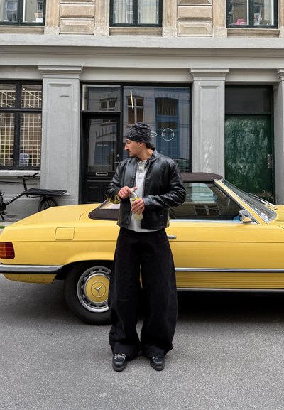 Chaqueta de cuero negra sobre camiseta gris, pantalones negros sueltos, sosteniendo un pulverizador. Coche descapotable amarillo con detalles cromados estacionado cerca.