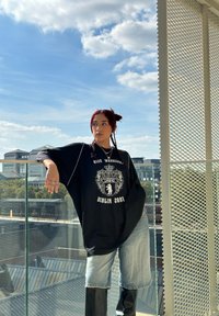 T-shirt oversized preto com estampado gráfico branco, combinado com calções de ganga azul claros e botas longas pretas contra um céu brilhante.