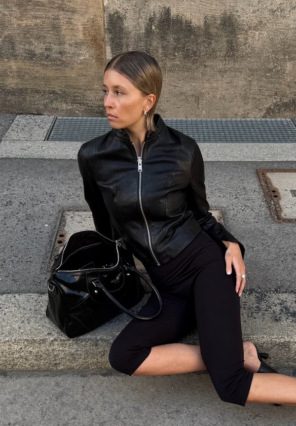 Jonge vrouw met naar achteren gekamde haren, gekleed in een zwart lederen jasje en zwarte cropped broek, zittend op een stoeprand naast een zwarte handtas.