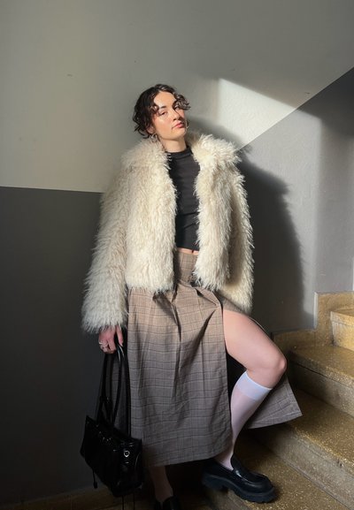 Chaqueta de piel sintética crema, top corto negro, falda midi de cuadros con abertura, calcetas blancas hasta la rodilla, bolso negro, zapatos negros gruesos, en interiores.