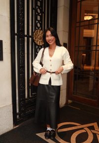 Mladá žena v bílém kardiganu, černé kožené sukni a mokasínech stojí u zdobné černo-zlaté brány a vstupu se skleněnými dveřmi.