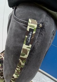 Grå denimbukser med en sidelomme med en camouflage-rem og et sort mærke med hvid tekst. Nærbillede af stofstrukturen.