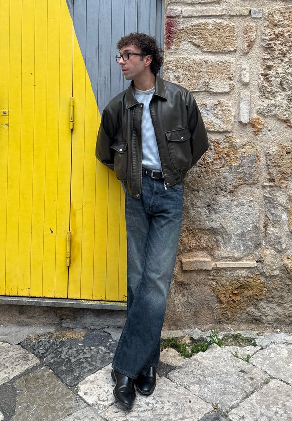 Άνδρας με σγουρά μαλλιά, γυαλιά, μαύρο δερμάτινο μπουφάν, λευκό πουλόβερ, σκούρο τζιν και μπότες στηρίζεται σε πέτρινο τοίχο δίπλα σε κίτρινες και γκρι ξύλινες πόρτες.