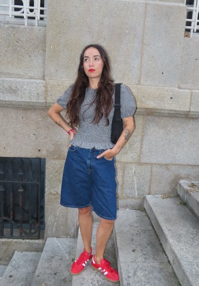 Top de manga corta a cuadros blanco y negro, pantalones cortos de mezclilla azules hasta la rodilla y zapatillas rojas con rayas blancas; de pie en unos escalones de piedra.