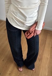 Kremowa bluzka z długim rękawem i ciemnoniebieskie dżinsy w typie dzwonów, w połączeniu z pogniecioną różową kopertówką. Beżowe sandały widoczne na stopach. Tło z drewnianej podłogi.