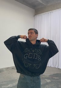 Black oversized hoodie with a textured design featuring the letters "GCDS" made of rhinestones, paired with high-waisted denim jeans.