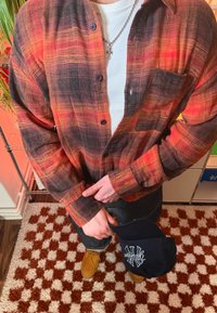 Narancssárga, piros és fekete kockás flanel ing, hosszú ujjú, kigombolatlan gallérral. Szürke bordázott alsóing, sötét farmernadrág és fekete sapka látható.