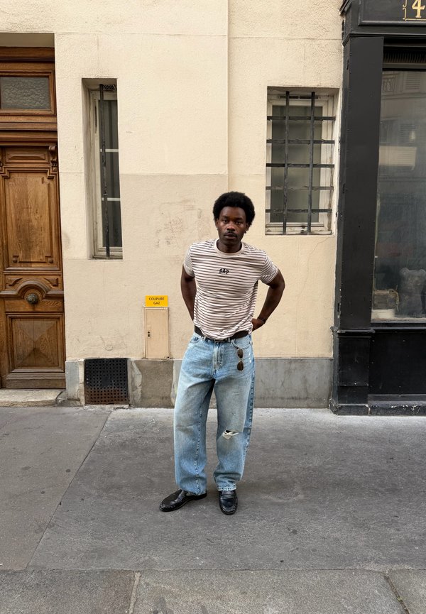 Hombre de pie en una acera de la ciudad con una camisa a rayas, vaqueros holgados rasgados con gafas de sol colgando y zapatos negros, junto a una pared beige.
