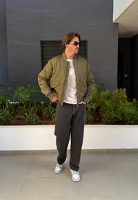 Olíva zöld bomber dzseki két elülső zsebbel, fehér póló, fekete bő szabású farmernadrág és szürke sportcipő. Egy szürke falnak támaszkodik, növények között.