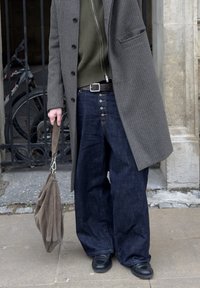 Ülisuurte tumedate teksapükstega kõrge vöökohaga, kombineeritud vormis oliivrohelise kudumiga ja ruudulise pika mantliga, hoides pruuni kotti.