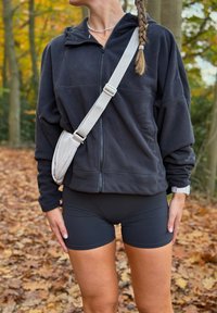 Svart fleecejakke med glidelås, lett stoff. Kombinert med tettsittende svarte shorts. Lysegrå crossbody-veske, naturlig utendørs bakgrunn med blader.