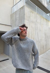 Grå och svart randig stickad tröja med hög krage, raglanärmar och en avslappnad passform, ihop med ljusblå jeans och glasögon.