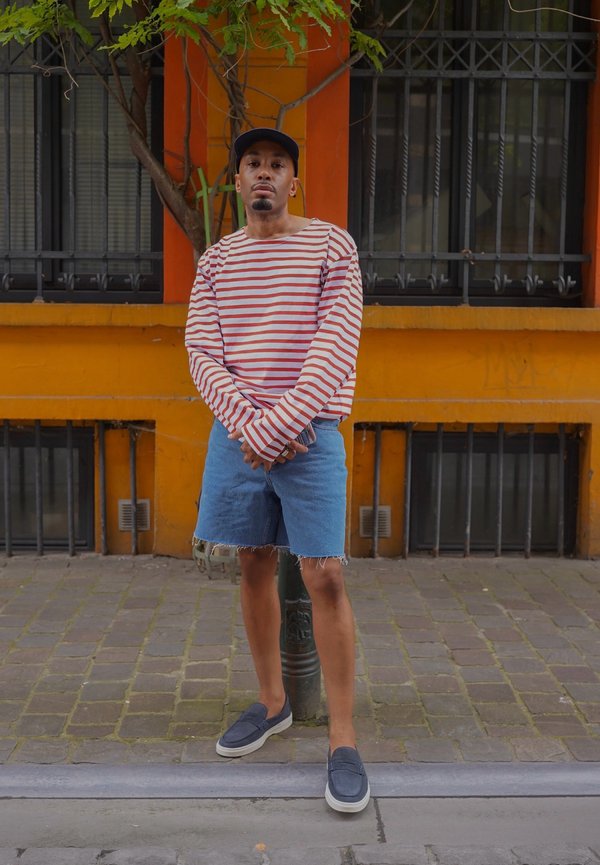 Uomo con camicia a righe a maniche lunghe, pantaloncini di denim e scarpe da ginnastica in piedi su una strada acciottolata davanti a un edificio arancione con finestre con sbarre.