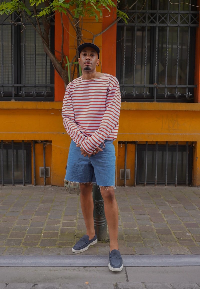 Homme en chemise rayée à manches longues, shorts en denim et baskets, debout dans une rue pavée devant un bâtiment orange avec des fenêtres grillagées.