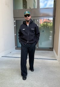 Bărbat îmbrăcat într-o hanorac negru, pantaloni negri, încălțăminte neagră, șapcă de baseball verde și ochelari de soare, stând în fața unor uși de sticlă.