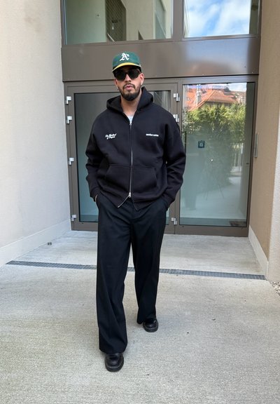 Hombre con sudadera negra, pantalones negros, zapatos negros, gorra de béisbol verde y gafas de sol de pie frente a puertas de cristal.