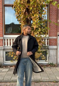 Sort ydre jakke med en struktureret grå sweater og lyseblå jeans. Lange ærmer og åben front. Murstensvæg og grønt løv i baggrunden.